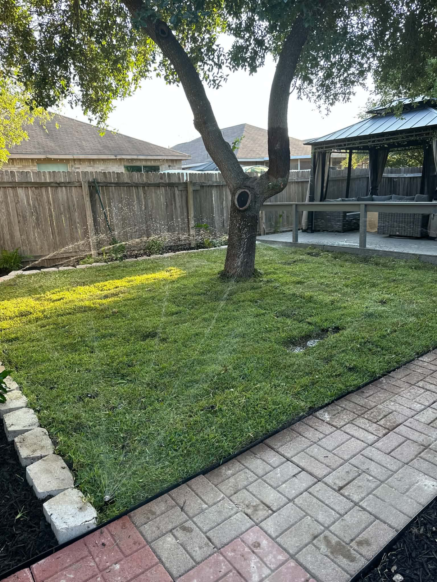Finished backyard lawn with sprinklers running beside a paved patio and gazebo